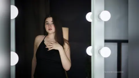 Young woman in a black halter top posing in a dimly lit room with vanity mirror lights.