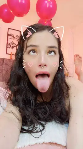 Playful young woman with cat ear filter and pink balloons in a cozy bedroom setting.