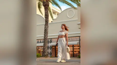 Stylish woman in white linen crop top and trousers walking near palm trees by luxury boutiques.