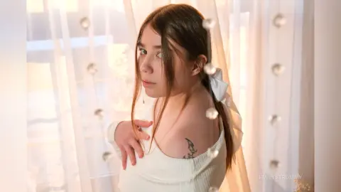 Young woman in a white ribbed top posing by sheer curtains in warm, soft sunlight.