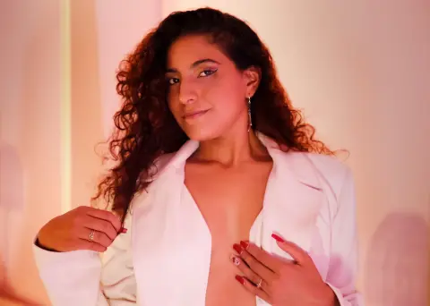 Young woman with curly hair wearing a white blazer in warm, soft lighting.