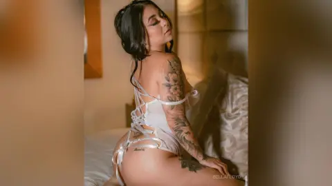 Tattooed woman in white lace lingerie posing on a bed in a warm, intimate bedroom setting.