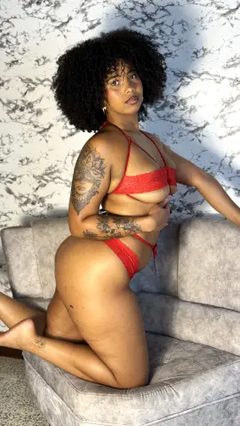 Curvy woman with afro hair in red lace lingerie posing on a grey sofa against a marble wall.
