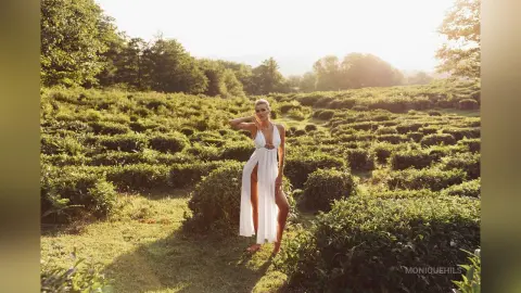 Elegant woman in a white flowing maxi dress posing in a sunlit tea plantation during golden hour.