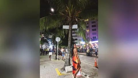 Stylish woman in a long orange dress walking on a tropical city street at night near palm trees.