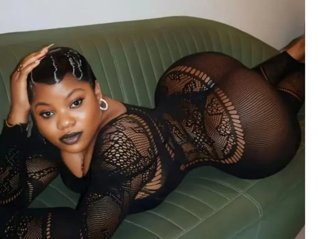 Curvy woman in a black lace bodysuit lounging on a green velvet sofa with a glamorous mood.