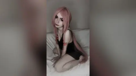 Young woman with long pink hair wearing a black tank top sitting on a white bed in a soft, dreamy mood.