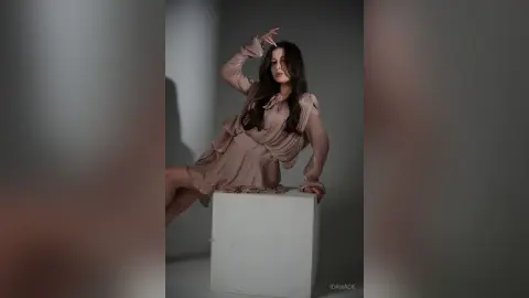 Elegant woman in a flowing mauve dress sitting on a white pedestal in a minimalist studio setting.