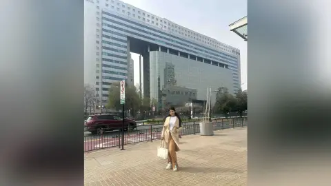 Stylish woman in a beige trench coat standing on a city sidewalk near a modern glass skyscraper.