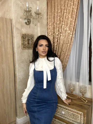 NoraSin poses in a white long-sleeve blouse with a neck tie and a tight blue denim dress, standing next to a wooden dresser and patterned curtains.