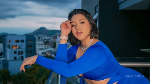 KimRoberth poses outdoors against a dark wall, wearing a blue long-sleeved top and a pearl necklace.