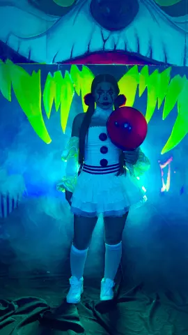 Girl in creepy clown costume holding a red balloon in a spooky, foggy blue-lit setting.