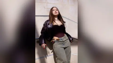 Stylish woman in a dark leather jacket and corset top posing confidently against a stone wall in sunlight.