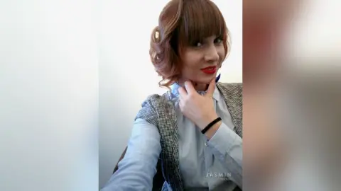 AryaSin poses with her hand under her chin, wearing a light blue button-down shirt and a grey patterned vest with her auburn hair styled in waves.