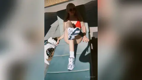 EllieFrosty sits on a light blue lounge chair wearing a red bikini top and white shorts, wearing sunglasses in the bright sunlight.
