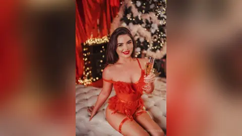Sofiya wears a red lace teddy while holding a glass of champagne on a white tufted bed.