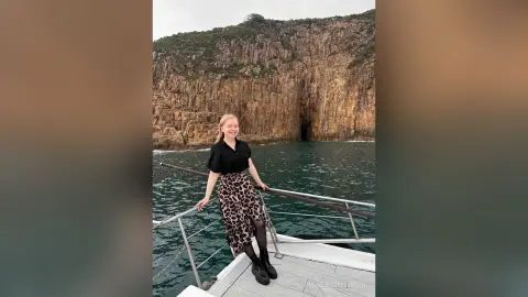 Smiling blonde woman in a black blouse and leopard print skirt relaxing on a boat near rocky ocean cliffs.