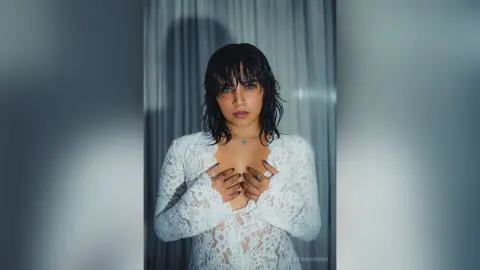 Moody portrait of a woman with dark hair in a white lace dress against a soft grey curtain background.
