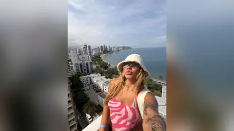 Blonde woman in a pink patterned top and white bucket hat enjoying a sunny coastal city view from a balcony.