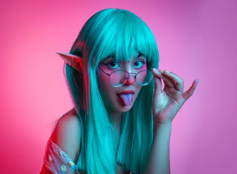 Cosplayer with teal hair and elf ears wearing glasses in a vibrant pink neon studio setting.
