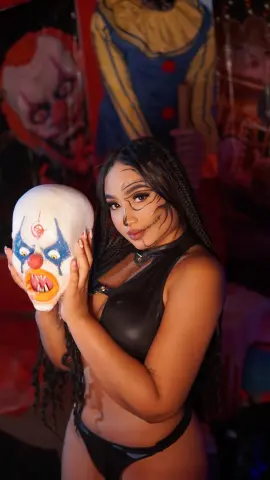 Woman in black cosplay outfit holding a creepy clown mask in a spooky, dark setting.
