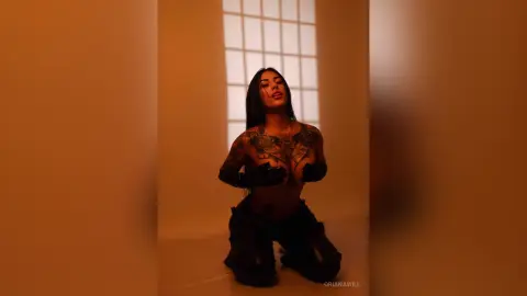 OrianaWill kneels in a room, showing her bare chest and tattoos while wearing black gloves and dark pants.