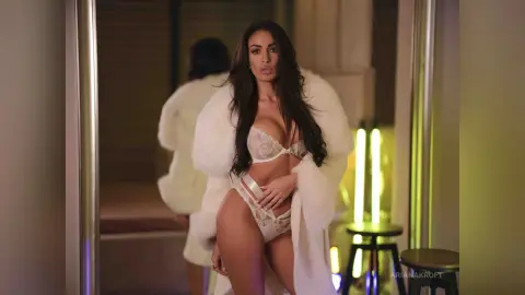 ArianaKroft wears a white furry robe and lace lingerie while posing in a dimly lit room.