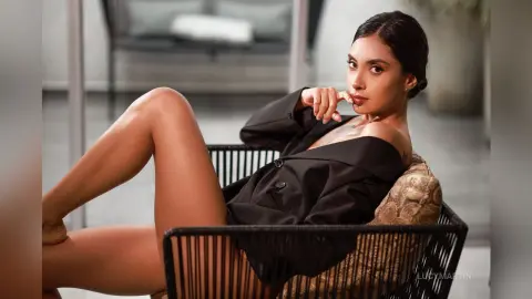 LucyMartin poses in a black blazer while sitting in a black wicker chair, showing off her bare legs and shoulders.