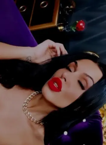 Glamorous woman with dark hair and red lipstick wearing a gold chain necklace in a moody, dark setting.