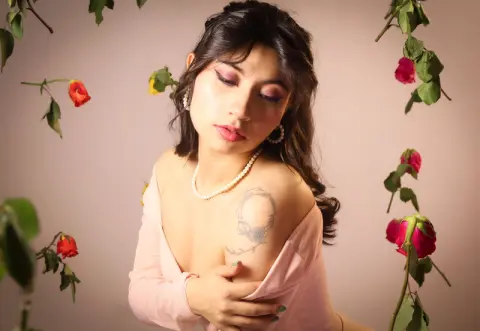 Young woman in a pink top and pearl necklace posing amidst rose vines in a soft, romantic mood.