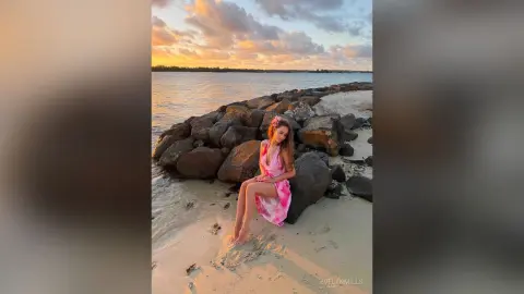 Young woman in a pink summer dress sitting on a rocky beach at sunset under a golden sky.