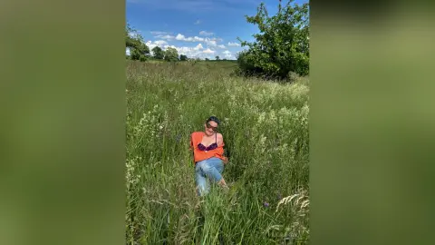 Smiling woman in an orange top and denim jeans relaxing in a sunny wildflower meadow.
