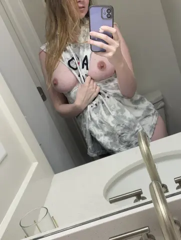 Young woman in a grey tie-dye oversized t-shirt taking a casual mirror selfie in a bright bathroom.