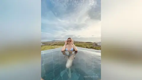 Woman in a white bikini relaxing in an infinity pool overlooking a scenic coastal landscape under a cloudy sky.