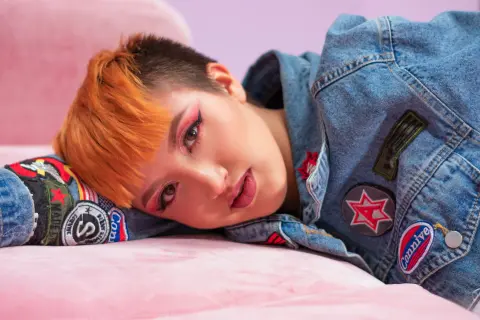 Woman with orange pixie cut in denim jacket lying on pink velvet sofa in soft pastel lighting.