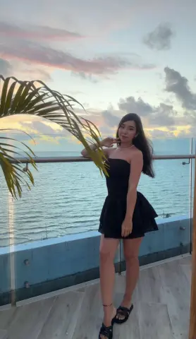 Young woman in a black mini dress posing on a balcony overlooking the ocean at sunset.