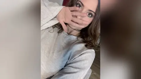 AriaLush covers her mouth with her hand, showing her eyes and long brown hair while wearing a light grey long-sleeve shirt.
