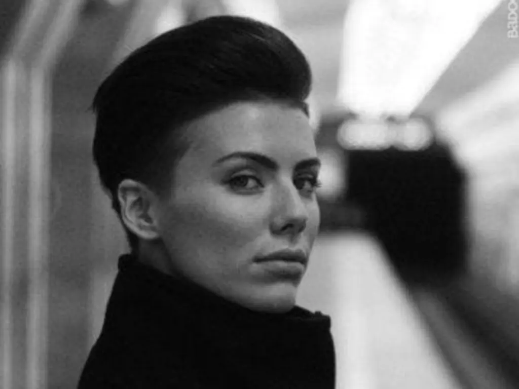 Moody black and white portrait of a woman with short dark hair wearing a black turtleneck in a blurred setting.