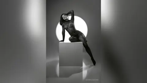 Elegant woman in a black lace bodysuit posing on a pedestal in a moody black and white studio setting.
