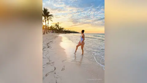 Woman in white swimsuit walking on a tropical beach at sunset with palm trees and ocean waves.