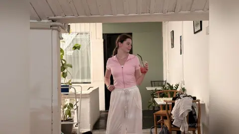 LiaMotzer stands in a bright room wearing a pink zip-up top and white trousers while holding a pink drink.