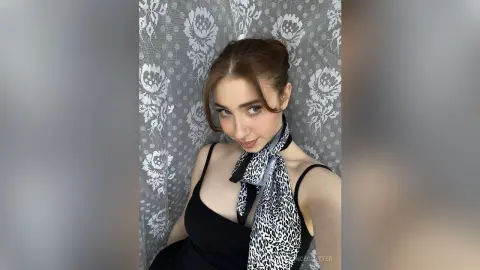 Young woman in a black dress and leopard print scarf posing against a grey floral lace curtain.