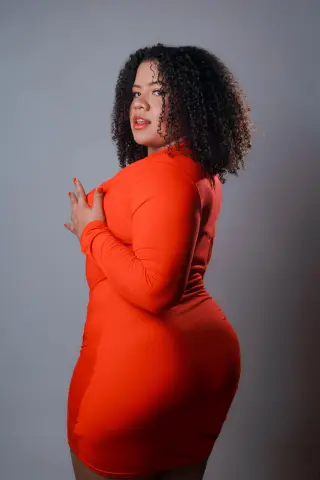 Curvy woman with curly hair wearing a bright orange bodycon dress posing against a grey studio background.