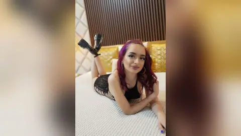 Smiling woman with purple hair in a black lace bodysuit relaxing on a white bed in a modern bedroom.