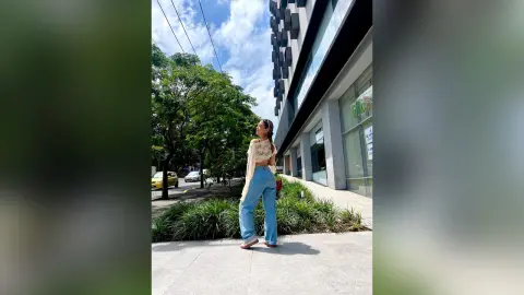 Stylish woman in a white crop top and blue jeans standing on a sunny city sidewalk near green trees.