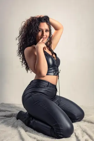 Curvy woman with long curly hair wearing a black leather corset and pants posing on a white rug.
