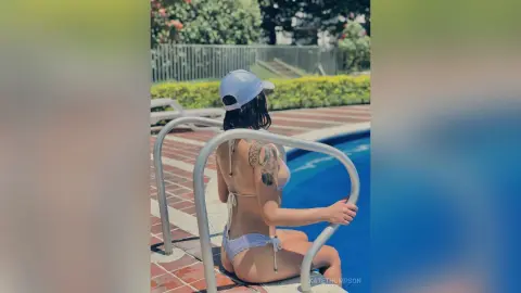 Woman in white bikini and light blue cap sitting by a sunny outdoor swimming pool.