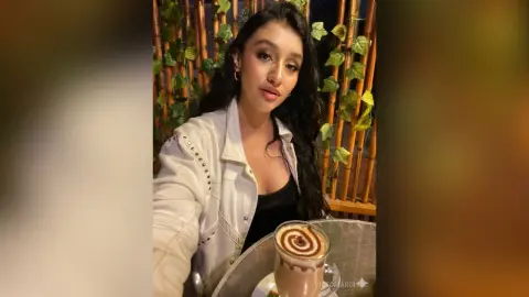 Young woman in a white denim jacket enjoying a latte at a cafe with lush green ivy background.