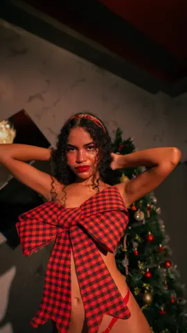 Young woman in a red checkered bow outfit posing by a festive Christmas tree in a moody indoor setting.