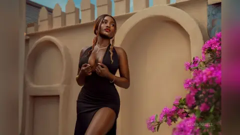 TinaRousell wears a low-cut black dress with a high slit, posing against a tan wall with bright pink flowers.